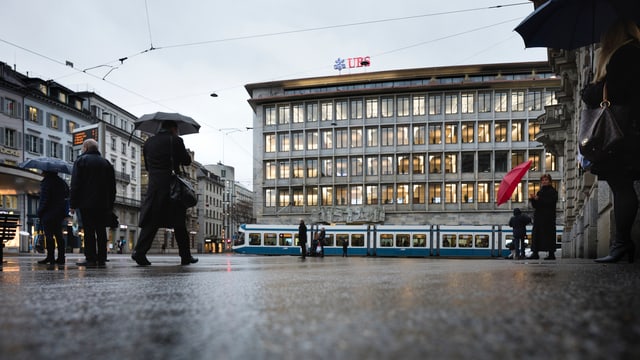 Leute stehen auf dem Zürcher Paradeplatz. Es regnet. Im Hintergrund das Gebäude der UBS. Leute stehen auf dem Zürcher Paradeplatz. Es regnet. Im Hintergrund das Gebäude der UBS.