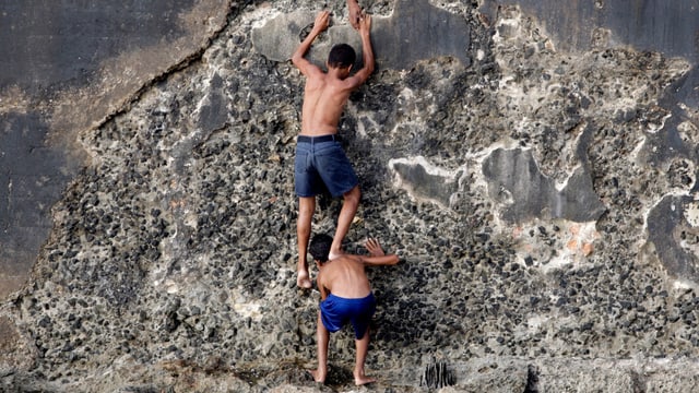 Zwei jungen stehen an einer Mauer, der eine bückt sich, der andere klettert über seine Schultern die Wand hoch.