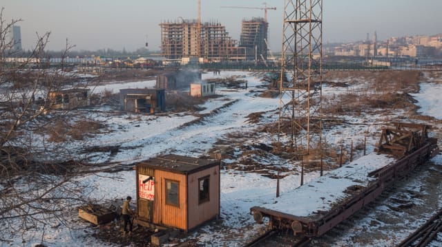 Man sieht von oben eine verschneite Brachlandschaft neben Bahngeleisen. Darauf mehrere kleine Baracken. Irgendwo steigt Rauch auf. Man sieht von oben eine verschneite Brachlandschaft neben Bahngeleisen. Darauf mehrere kleine Baracken. Irgendwo steigt Rauch auf.