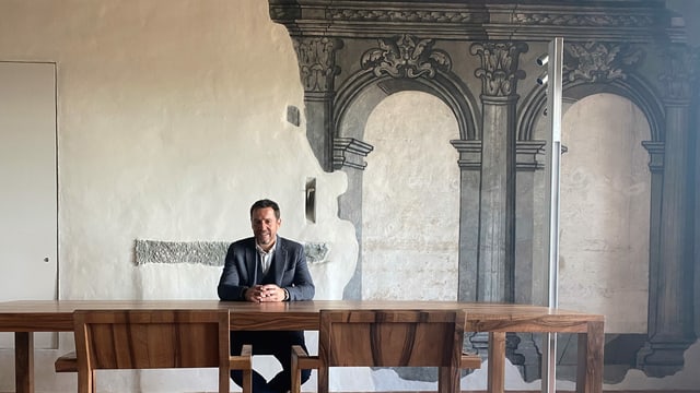 Er sitzt an einem Holztisch, dahinter eine weisse Wand, auf der in grau Rundbögen und Säulen gemalt sind. Er sitzt an einem Holztisch, dahinter eine weisse Wand, auf der in grau Rundbögen und Säulen gemalt sind.
