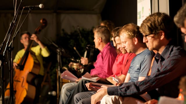 Christian Brantschen spielt im Hintergrund Kontrabass, im Vordergrund sitzen verschiedene Mitglieder der Gruppe «Bern ist überall»..