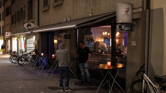 Bar an einer gepflasterten Altstadtgasse. Draussen ist es dunkel, zwei Männer stehen vor der BAr. Durch das Fenster sieht man Menschen im beleuchteten Innenraum. Bar an einer gepflasterten Altstadtgasse. Draussen ist es dunkel, zwei Männer stehen vor der BAr. Durch das Fenster sieht man Menschen im beleuchteten Innenraum.