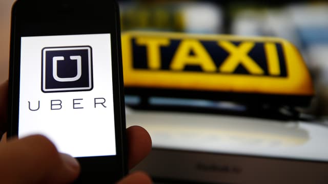 Eine Hand hält ein Smartphone mit der Uber-App vor einem Taxi. Eine Hand hält ein Smartphone mit der Uber-App vor einem Taxi.