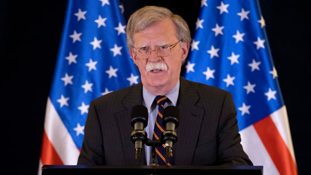 John Bolton spricht in Mikrofone, hinter ihm zwei USA-Flaggen. John Bolton spricht in Mikrofone, hinter ihm zwei USA-Flaggen.