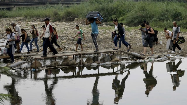 Eine Gruppe Flüchtlinge überquert mitsamt Koffern und Taschen via Holzsteg einen Fluss. Eine Gruppe Flüchtlinge überquert mitsamt Koffern und Taschen via Holzsteg einen Fluss.