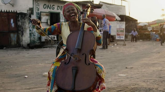 Eine afrikanische Frau spielt ein Cello auf einer Naturstrasse.
