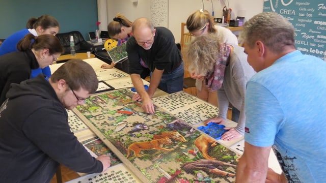 Acht Leute bücken sich über das Puzzle auf einem langen Tisch. Acht Leute bücken sich über das Puzzle auf einem langen Tisch.