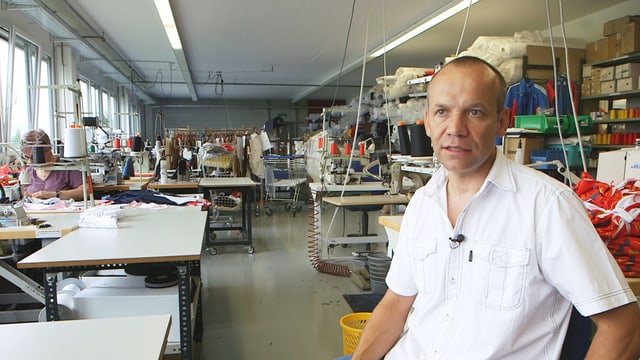 Walter Graf sitzt imn der Näherei, im Hintergrund sieht man Leute an den Nähmaschinen arbeiten. Walter Graf sitzt imn der Näherei, im Hintergrund sieht man Leute an den Nähmaschinen arbeiten.