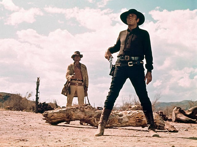 Filmszene: Ein Mann in Schwarzer Kleidung mit Cowboy-Hut greift nach seinem Revolver. Hinter ihm steht ein anderer Mann, der seine Pistole auf ihn richtet. Filmszene: Ein Mann in Schwarzer Kleidung mit Cowboy-Hut greift nach seinem Revolver. Hinter ihm steht ein anderer Mann, der seine Pistole auf ihn richtet.