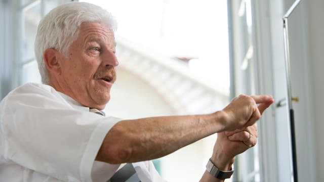 Ein älterer Herr gestikuliert mit seinen Händen vor einem offenen Fenster. Ein älterer Herr gestikuliert mit seinen Händen vor einem offenen Fenster.