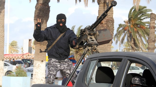 Ein Mann in Tarnhosen und schwarzer Gesichtsmaske steht auf einem Pickup an einem Maschinengewehr und macht das Victory-Zeichen.