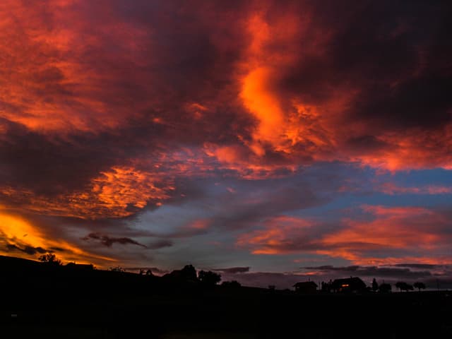 Roter, gelber und hellblauer Himmel mit Wolken. Unten die Konturen von Bäumen und Bergen.