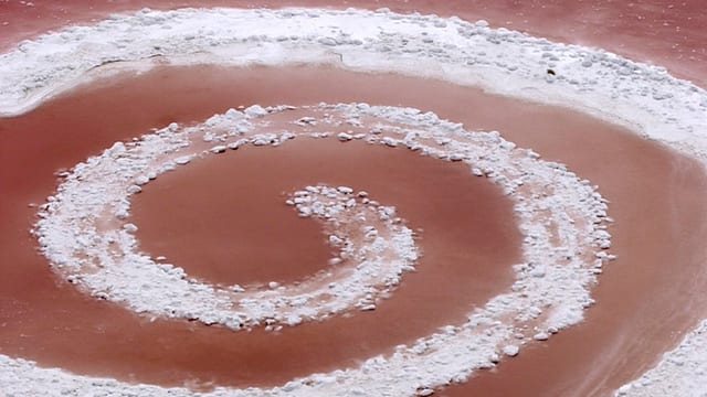 Durch das Wasser eines brau-rot schimmernden Sees zieht eine Salzspur einen mehrfach gewickelten Kringel. Durch das Wasser eines brau-rot schimmernden Sees zieht eine Salzspur einen mehrfach gewickelten Kringel.