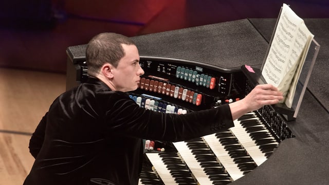 Ein Mann mit kurzgeschnittenen Haaren sitzt an einer Orgel und blättert in der Partitur. Ein Mann mit kurzgeschnittenen Haaren sitzt an einer Orgel und blättert in der Partitur.