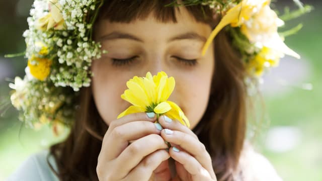 Ein Mädchen mit Blumenkranz auf dem Kopf riecht an einer gelben Blume. 