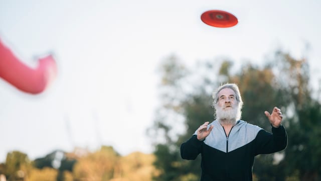 Ein Alter Mann mit graunen Haaren und Bart steht in einem Park und fängt einen Frisbee. Ein Alter Mann mit graunen Haaren und Bart steht in einem Park und fängt einen Frisbee.