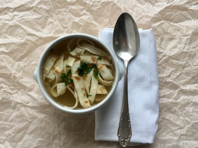 Eine Suppenschüssel mit Hühnerbouillon und Flädli. Daneben ein silberner Suppenlöffel. Eine Suppenschüssel mit Hühnerbouillon und Flädli. Daneben ein silberner Suppenlöffel.