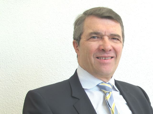 Ueli Graf in blau-gelb gestreifter Krawatte, blauem Hemd und schwarzem Jacket sitzt an einem runden Holztisch. Ueli Graf in blau-gelb gestreifter Krawatte, blauem Hemd und schwarzem Jacket sitzt an einem runden Holztisch.