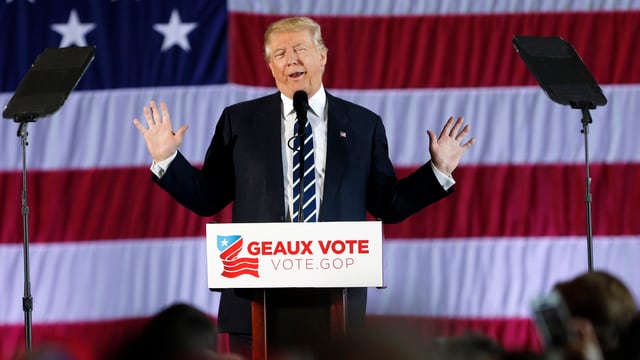 Trump spicht vor einer riesigen US-Flagge auf einer Bühne in ein Mikrofon, die Arme hat er seitlich erhoben. Trump spicht vor einer riesigen US-Flagge auf einer Bühne in ein Mikrofon, die Arme hat er seitlich erhoben.
