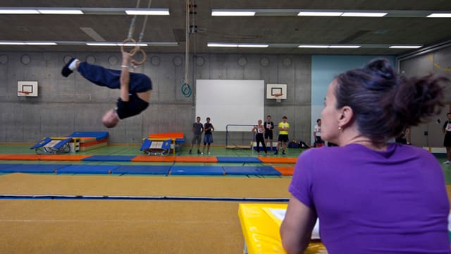 Eine Turnlehrerin beobachtet einen Schüler der an Ringen hängt. Eine Turnlehrerin beobachtet einen Schüler der an Ringen hängt.