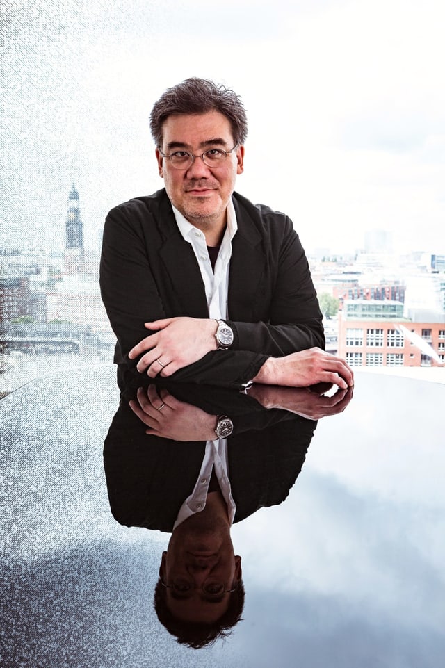 Ein Mann mittleren Alters lehnt sich im Anzug über einen Glastisch. Dieser spiegelt ihn. Im Hintergrund ist eine Stadt zu sehen. Ein Mann mittleren Alters lehnt sich im Anzug über einen Glastisch. Dieser spiegelt ihn. Im Hintergrund ist eine Stadt zu sehen.