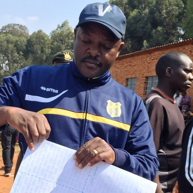 Ein Afrikaner in blaubem Trainingsanzug und Baseball-Mütze mit einem Bogen weissen Papiers in der Hand, um ihn herum stehen weitere Menschen. Ein Afrikaner in blaubem Trainingsanzug und Baseball-Mütze mit einem Bogen weissen Papiers in der Hand, um ihn herum stehen weitere Menschen.