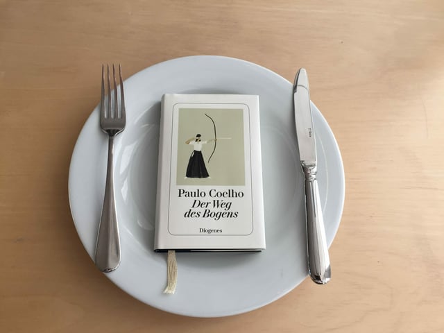 Das Buch «Der Weg des Bogens» von Paulo Coelho liegt auf einem weissen Teller. Messer und Gabel liegen auf dem Teller daneben. 
