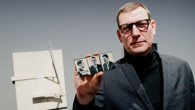 Ein Mann steht vor einem abstrakten Relief. Er hält schwarz-weisse Porträtfotos in der Hand. Ein Mann steht vor einem abstrakten Relief. Er hält schwarz-weisse Porträtfotos in der Hand.