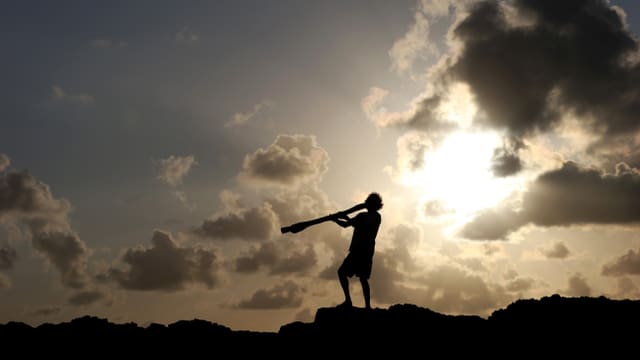 Australischer Ureinwohner auf einem Felsen im Gegenlicht, er spielt Didgeridoo. Australischer Ureinwohner auf einem Felsen im Gegenlicht, er spielt Didgeridoo.
