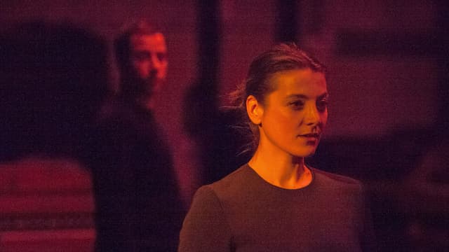 Eine Frau steht auf der Bühne. Hinter ihr steht ein Mann. Sie sind rot beleuchtet. Eine Frau steht auf der Bühne. Hinter ihr steht ein Mann. Sie sind rot beleuchtet.