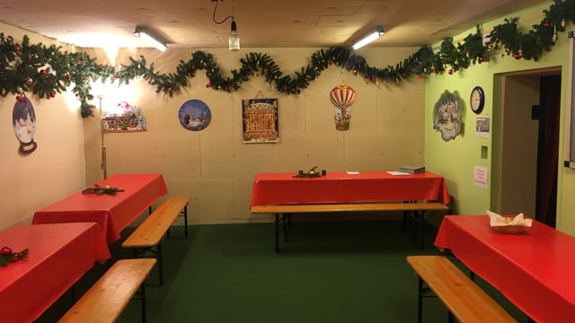 Ein Zivilschutzraum mit vier roten Tischen, bedeckt mit roten Tischtüchern. An der Wand Tannenzweige, geschmückt mit Christbaumkugeln. Ein Zivilschutzraum mit vier roten Tischen, bedeckt mit roten Tischtüchern. An der Wand Tannenzweige, geschmückt mit Christbaumkugeln.