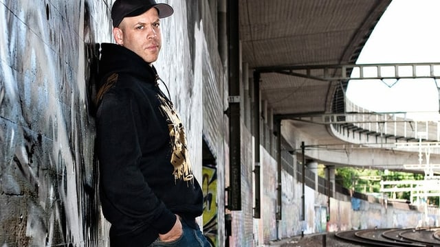 Mann mit Baseball-Mütze lehnt neben einem Bahngleis an einer besprayten Wand Mann mit Baseball-Mütze lehnt neben einem Bahngleis an einer besprayten Wand