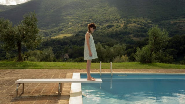 Eine junge Frau steht in einem Sommerkleid auf einem Pool-Sprungbrett vor einer hügelig-ländlichen Kulisse. Eine junge Frau steht in einem Sommerkleid auf einem Pool-Sprungbrett vor einer hügelig-ländlichen Kulisse.