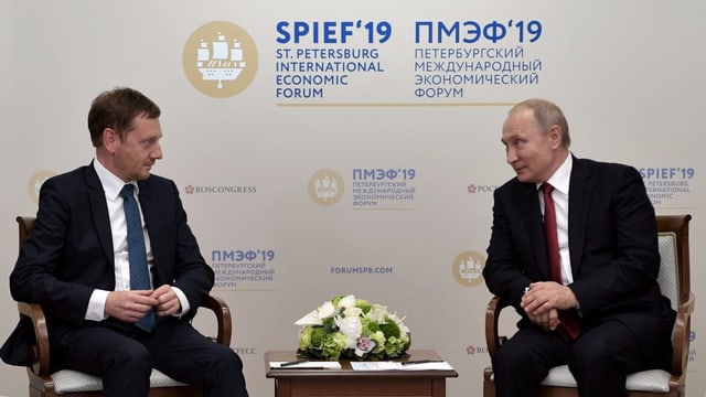 Zwei Männer sitzen sich gegenüber. Dahinter eine Schrift: St. Petersburg International Economic Forum. Zwei Männer sitzen sich gegenüber. Dahinter eine Schrift: St. Petersburg International Economic Forum.