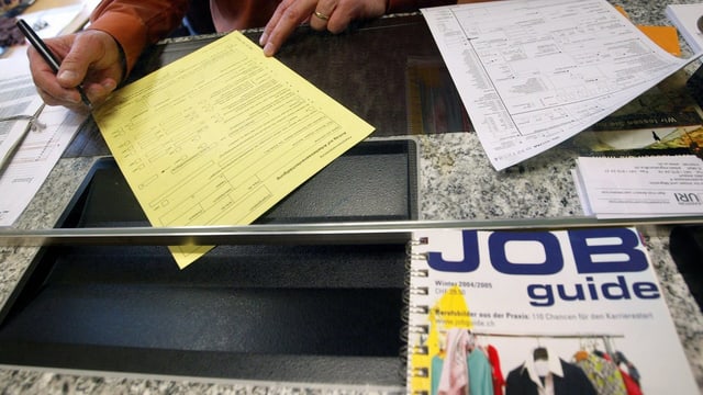 Eine Person am Schalter. Vor dem Schalter ist eine Brochure mit dem Titel «Job Guide» Eine Person am Schalter. Vor dem Schalter ist eine Brochure mit dem Titel «Job Guide»