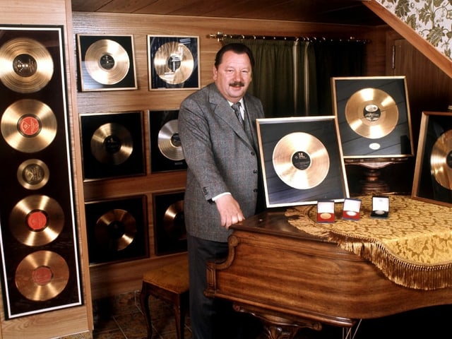 Ein Mann steht in einem Raum voller Goldener Schallplatten vor einem Flügel. Ein Mann steht in einem Raum voller Goldener Schallplatten vor einem Flügel.