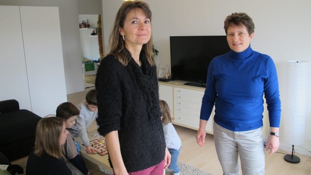 Zwei Frauen blicken in die Kamera, im Hintergrund Kinder und eine Frau am spielen. Zwei Frauen blicken in die Kamera, im Hintergrund Kinder und eine Frau am spielen.