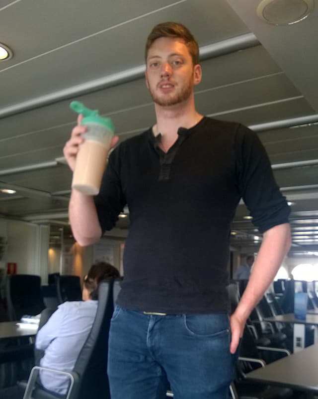 Ein Mann mit schwarzem T-Shirt. In seiner Hand ein grosser Schüttelbecher, darin eine hellbraune Flüssigkeit. Ein Mann mit schwarzem T-Shirt. In seiner Hand ein grosser Schüttelbecher, darin eine hellbraune Flüssigkeit.
