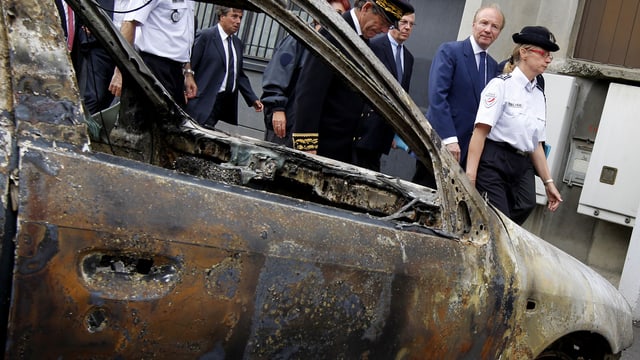 Ein ausgebranntes Auto, dahinter Polizei und Politiker.