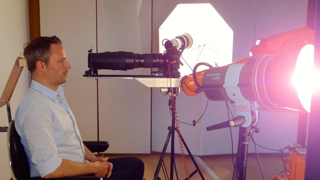 Daniel Wagner sitzt auf einem Stuhl, wird von allen Seiten beleuchtet - vor ihm der Roboterarm mit der darauf montierten Kamera. Daniel Wagner sitzt auf einem Stuhl, wird von allen Seiten beleuchtet - vor ihm der Roboterarm mit der darauf montierten Kamera.