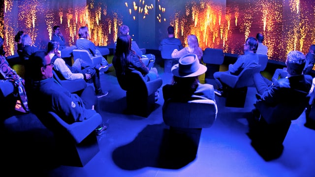 Leute sitzen in einem blau beleuchteten Museumssaal un dsehen sich feurige Animationen an. Leute sitzen in einem blau beleuchteten Museumssaal un dsehen sich feurige Animationen an.