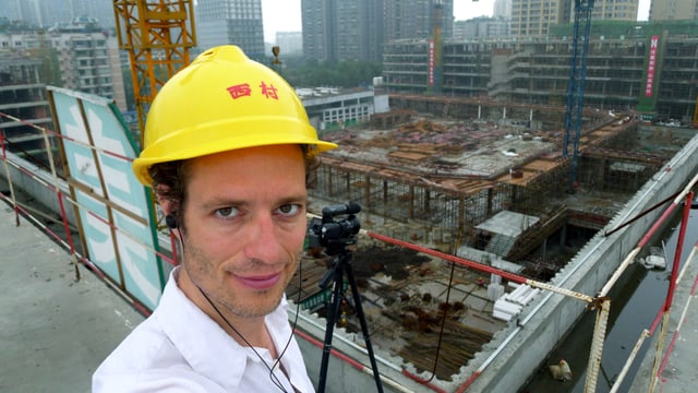 Stefan Kaegi mit gelbem Helm und Videokamera – mit Sicht auf entstehende Gebäude. Stefan Kaegi mit gelbem Helm und Videokamera – mit Sicht auf entstehende Gebäude.