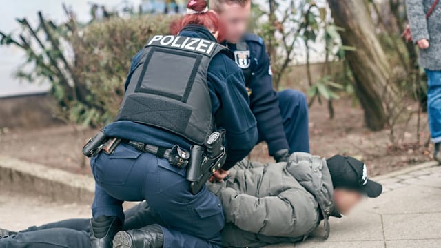 Eine Polizistin und ein Polizist drücken einen Mann auf den Boden und halten seine Arme auf den Rücken. Eine Polizistin und ein Polizist drücken einen Mann auf den Boden und halten seine Arme auf den Rücken.