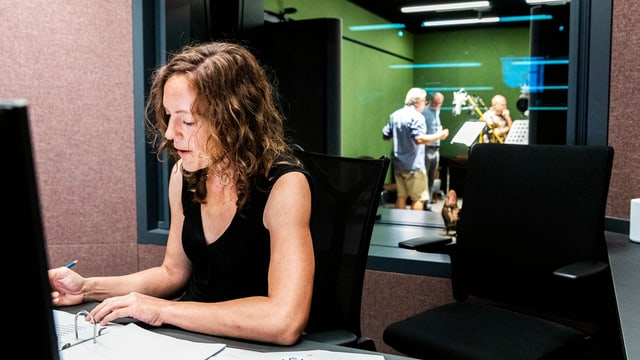 Ein Frau sitzt in einem Raum vor einem Zettel. Hinter ihr sieht man durch ein Fenster in ein Radiostudio. Ein Frau sitzt in einem Raum vor einem Zettel. Hinter ihr sieht man durch ein Fenster in ein Radiostudio.