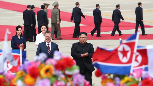 Moon und Kim mit ihren Frauen auf rotem Teppich, im Vordergrund Fahnen und Blumen. Moon und Kim mit ihren Frauen auf rotem Teppich, im Vordergrund Fahnen und Blumen.