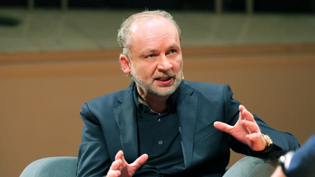 Ein Mann im Anzug sitzt auf einem Sessel, spricht und gestikuliert. Ein Mann im Anzug sitzt auf einem Sessel, spricht und gestikuliert.