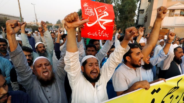 Protestierende Männer mit Bart schreiben Parolen, einer hält ein rotes Transparent mit pakistanischen Schriftzeichen in die Höhe. Protestierende Männer mit Bart schreiben Parolen, einer hält ein rotes Transparent mit pakistanischen Schriftzeichen in die Höhe.