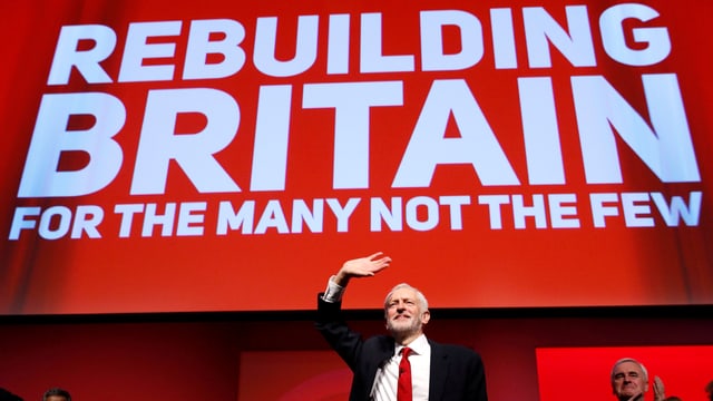 Labour-Chef Corbyn klein unter einem roten Banner mit der weissen Aufschrift "Rebuilding Britain for the many not the fews". Labour-Chef Corbyn klein unter einem roten Banner mit der weissen Aufschrift "Rebuilding Britain for the many not the fews".