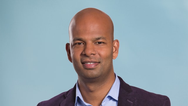 Rupan Sivaganesan steht Portrait vor einem blauen Hintergrund. Rupan Sivaganesan steht Portrait vor einem blauen Hintergrund.