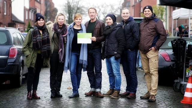 Sieben Personen stehen nebeneinander und schauen in die Kamera. Einer hält einen grünen Zettel. Sieben Personen stehen nebeneinander und schauen in die Kamera. Einer hält einen grünen Zettel.